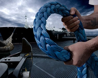 Foto de dos manos sujetando una cuerda gruesa. En el fondo hay un barco en el mar.